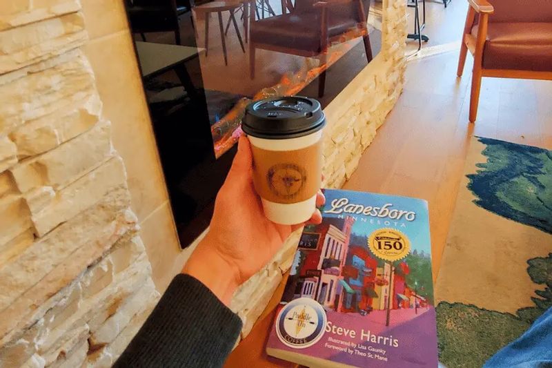 Hand holding a coffee near a fireplace with the book Lanesboro, Minnesota by Steve Harris on a table in a cozy indoor setting.