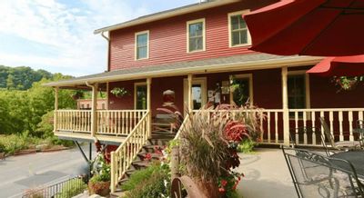 The Root River Inn features a red historic building with a wraparound porch, outdoor seating, and landscaped planters, overlooking the Root River Valley on a sunny day in Lanesboro.