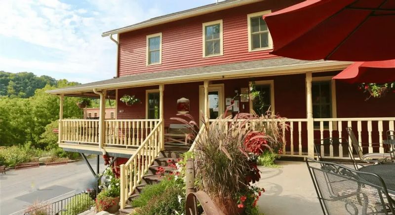 The Root River Inn features a red historic building with a wraparound porch, outdoor seating, and landscaped planters, overlooking the Root River Valley on a sunny day in Lanesboro.