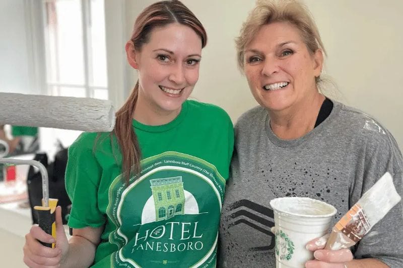 Two women smile at the camera while holding painting supplies during an interior renovation, one wearing a green Hotel Lanesboro T-shirt and holding a paint roller, the other holding a paint cup and brush.