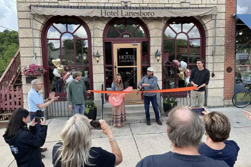 A ribbon-cutting ceremony takes place outside Hotel Lanesboro, with several people holding an orange ribbon in front of the historic arched entrance while a small crowd gathers and takes photos on the sidewalk.