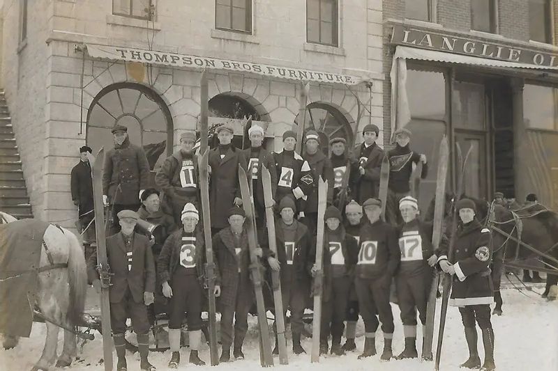 A historic black-and-white photograph shows a group of men posing with long wooden skis in front of the Thompson Bros. Furniture storefront in winter, with snow on the ground and period clothing indicating an early 20th-century scene.