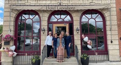 A group of women stand smiling on the front steps of Hotel Lanesboro, framed by arched windows and the hotel entrance, with decorative planters and heart-shaped balloons adding a celebratory touch.