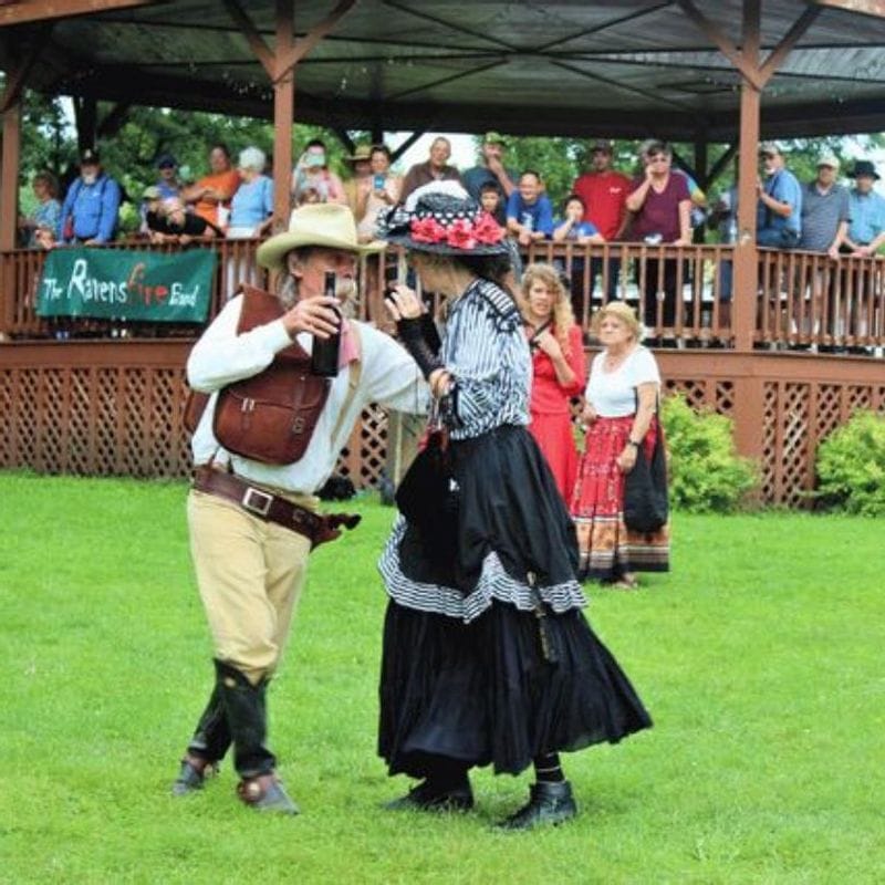 Performers in Old West–style costumes dance on the grass at Sylvan Park during Buffalo Bill Days, with spectators watching from the gazebo in the background.