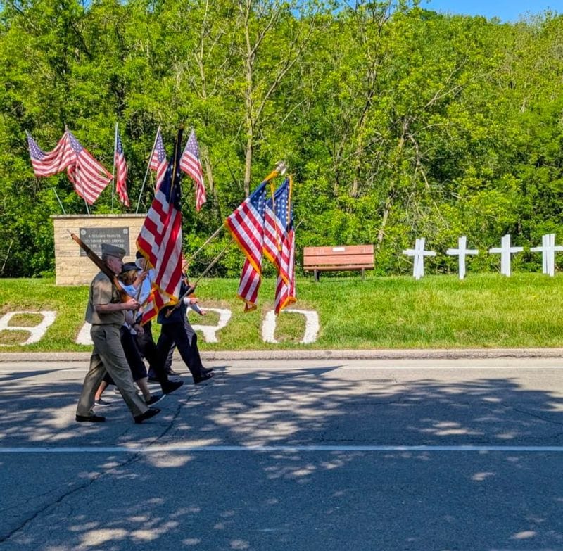 Color guard marching with American flags during the Lanesboro Memorial Day parade, passing a veterans memorial with white crosses and patriotic displays in the background.