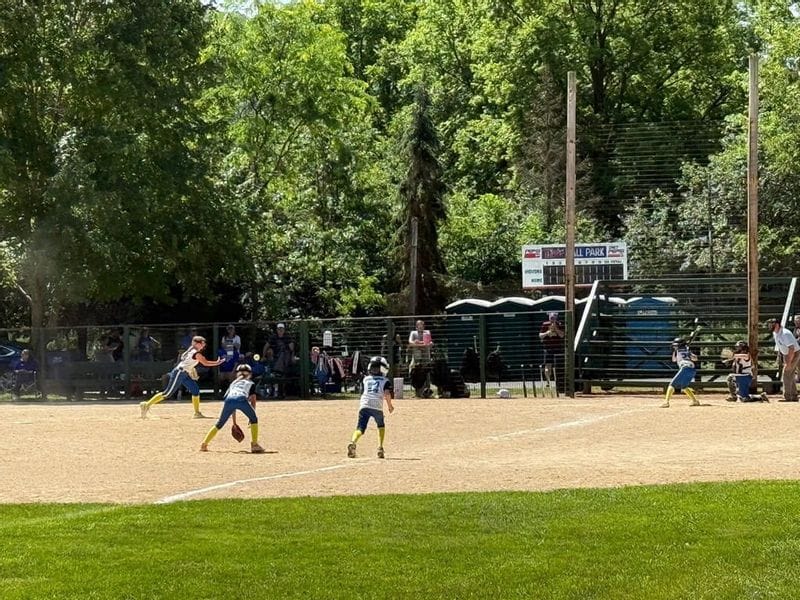 Girls fastpitch softball game in progress at a Whalan ballpark, with players fielding a hit on the infield while spectators watch from the bleachers and dugout area in the background.