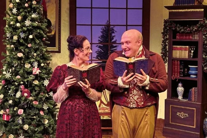 Two actors in period costumes perform onstage during a holiday production at the Commonweal Theatre in Lanesboro, standing beside a decorated Christmas tree and holding open books.