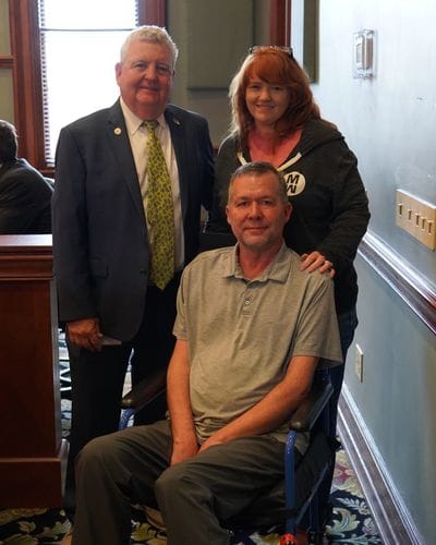 Mayor Doug Hendrix stands alongside Scott Bell and a companion during a City Council meeting as the City of Statesville proclaims May 2026 as ALS Awareness Month. Scott Bell, seated in a wheelchair, smiles toward the camera while Mayor Hendrix stands beside him in a dark suit and patterned tie. The group poses together inside the council chambers following the proclamation presentation.