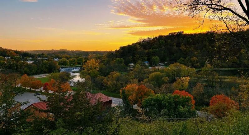Fall sunset view from Church Hill Overlook in Lanesboro, Minnesota, looking down over the Root River Valley with colorful autumn trees, the river winding past the dam, and wooded bluffs glowing under a golden sky.