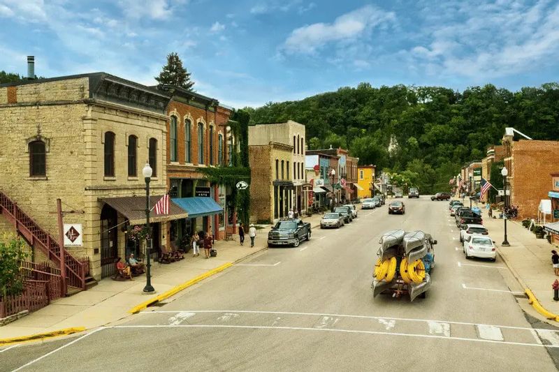 Downtown Lanesboro, Minnesota, with historic brick storefronts lining Main Street as a vehicle carrying yellow river tubes drives through town on a summer day, wooded bluffs rising in the background.