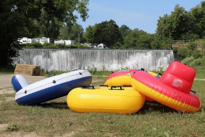 Colorful river tubes resting on the grass near the Lanesboro dam along the Root River, with the waterfall visible in the background on a sunny summer day.