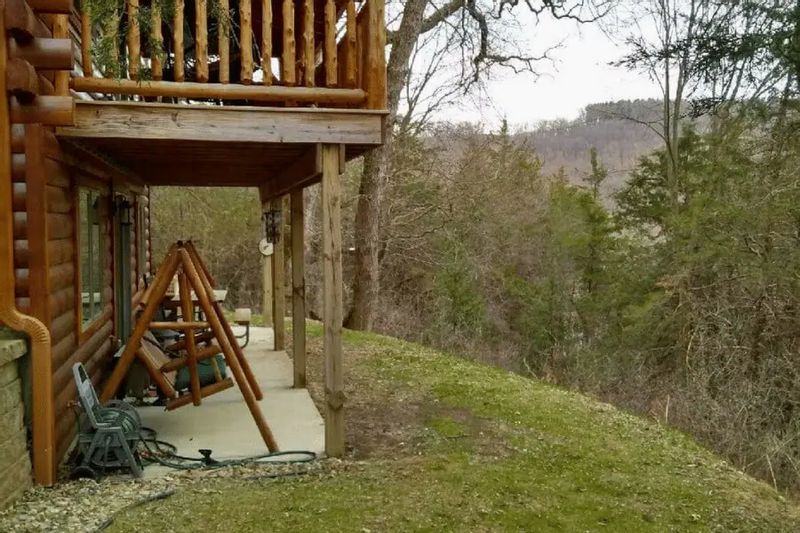 A covered porch at Root River Valley Cabin features a wooden swing and log railing, overlooking a wooded hillside and scenic valley views in a quiet natural setting.