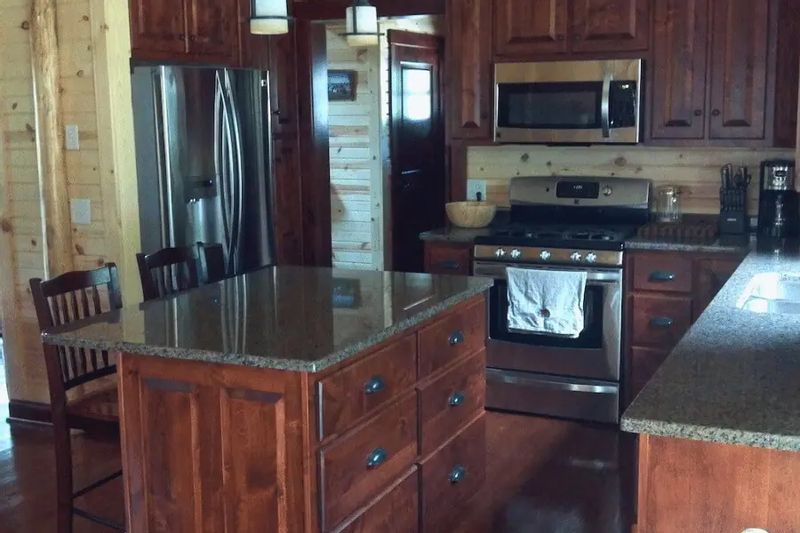 The rustic kitchen at Root River Valley Cabin features wood-paneled walls, a central island with granite countertop, and modern stainless steel appliances including a refrigerator, stove, and microwave.