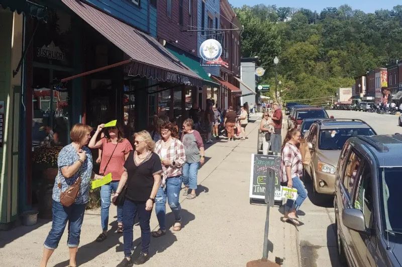Visitors stroll along Main Street in downtown Lanesboro, Minnesota, browsing local shops and restaurants on a sunny day, with parked cars and wooded bluffs rising in the background.