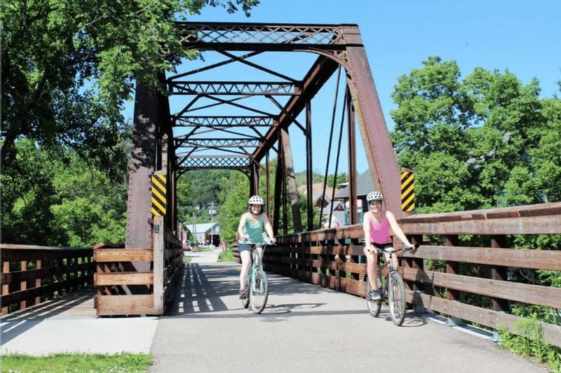 Two women ride bicycles across a historic bridge on the Root River Trail in Lanesboro, surrounded by summer greenery.