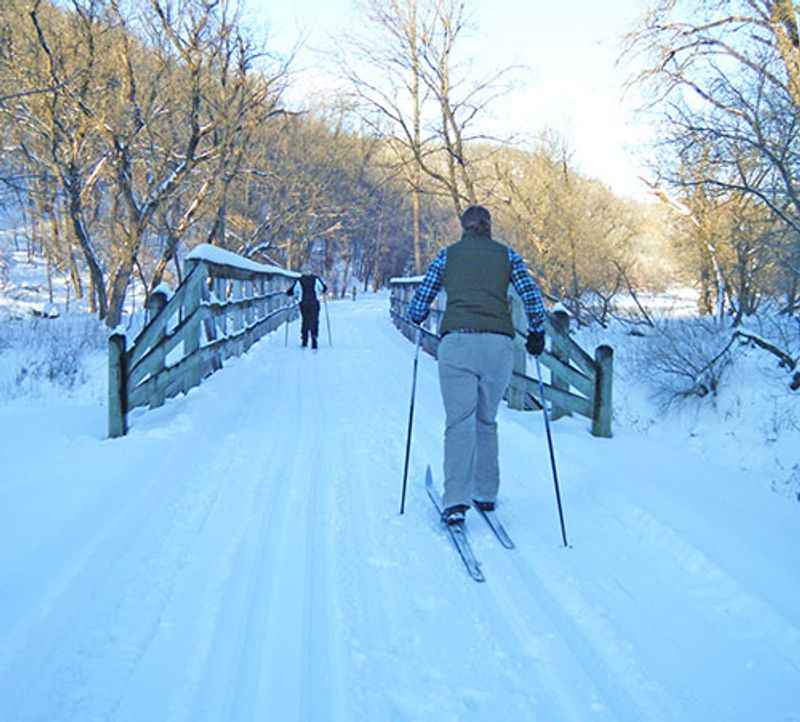 Cross-country skiers gliding across a snow-covered bridge on the Root River Trail during winter in Lanesboro, Minnesota.