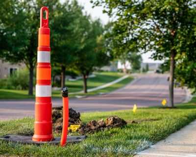 An orange utility marker and exposed service line stand beside a neighborhood sidewalk, indicating underground utility or infrastructure work in progress. A quiet residential street lined with trees and well-kept lawns stretches into the distance on a sunny day.