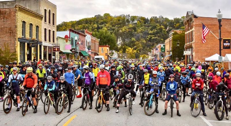 Hundreds of cyclists line up on Main Street in downtown Lanesboro for the Filthy 50 gravel bike race, with historic brick buildings and fall-colored bluffs in the background.