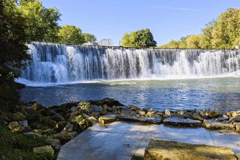 Wide view of water flowing over the Lanesboro dam into the Root River, with trees along the riverbank.