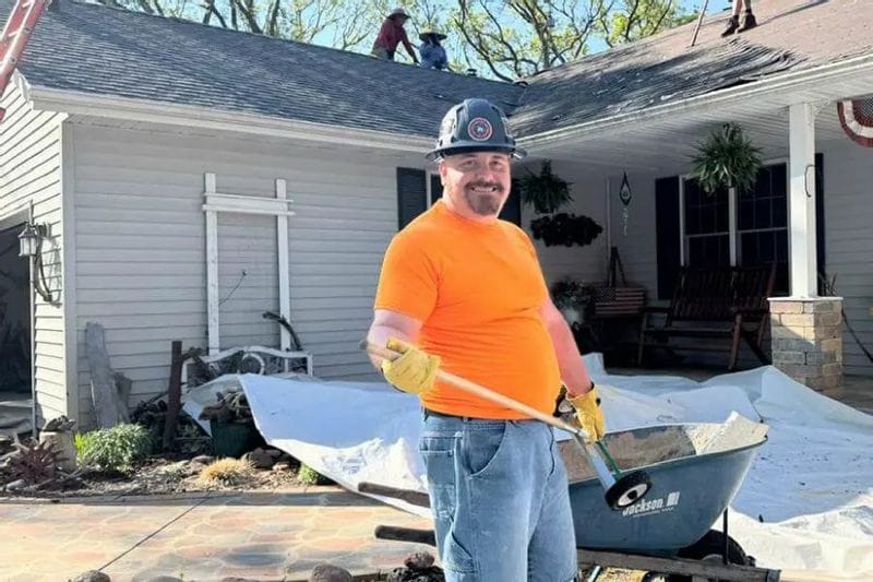 A Minnesota Building Contractors employee smiles while holding a shovel in front of a home undergoing roof work, with additional crew members visible working on the roof behind him.