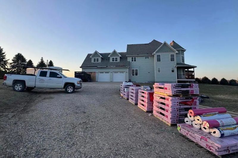 Stacks of roofing materials sit in a driveway beside a rural home as a Minnesota Building Contractors truck is parked nearby, ready for a roof replacement project.