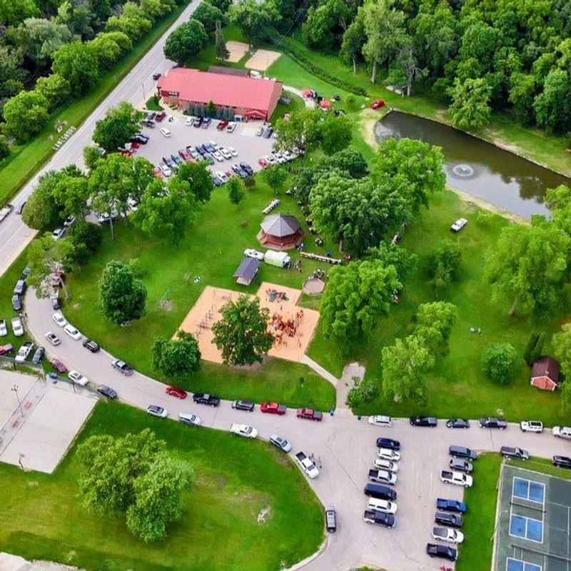 Aerial view of Lanesboro's Sylvan Park, showing cars parked all along the streets and parking lots, as well as the community center, gazebo, playground, pond, picnic shelters, and basketball courts all surrounded by lush summer greenery