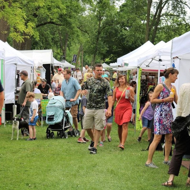 Crowds stroll through Art in the Park in Lanesboro, browsing white-tent artist booths under leafy trees, with families, children, and a stroller moving along the grassy park pathway during the outdoor arts festival.