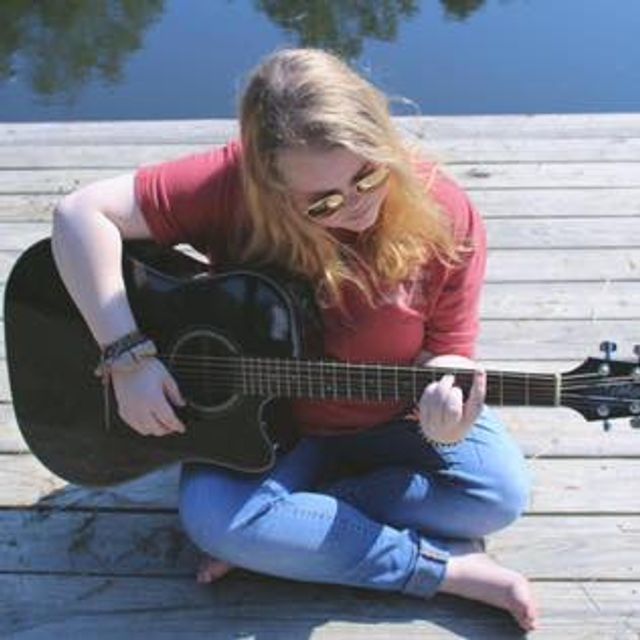 Musician Hailey Holt sits cross-legged on a wooden dock beside the water while playing a black acoustic guitar. She wears sunglasses, a red shirt, and jeans as sunlight reflects off the water behind her. The peaceful outdoor setting creates a calm and artistic atmosphere centered around live music.