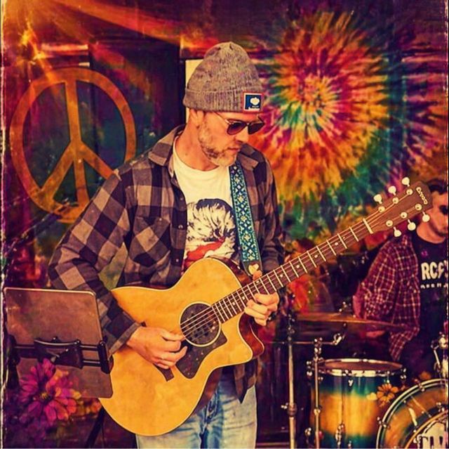 Josh Perryman performs onstage while playing an acoustic guitar in front of a colorful tie-dye backdrop featuring a peace sign. Wearing a knit beanie, sunglasses, and a plaid flannel shirt, the performer stands under warm lighting as a drummer plays in the background. The vibrant setting creates a relaxed, festival-style atmosphere.