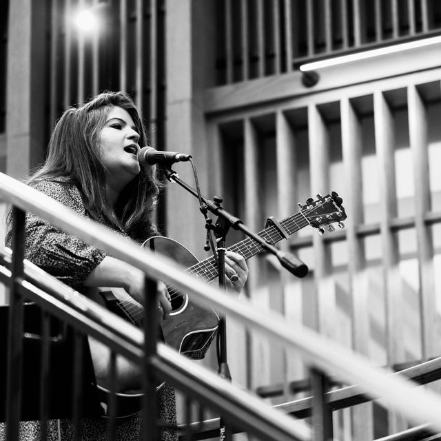 In a black-and-white photo, musician Karla Causey performs live while singing into a microphone and playing an acoustic guitar. She stands on a stage or elevated platform surrounded by railings and architectural details in the background. The image captures an expressive performance moment with dramatic lighting and a focus on live music artistry.