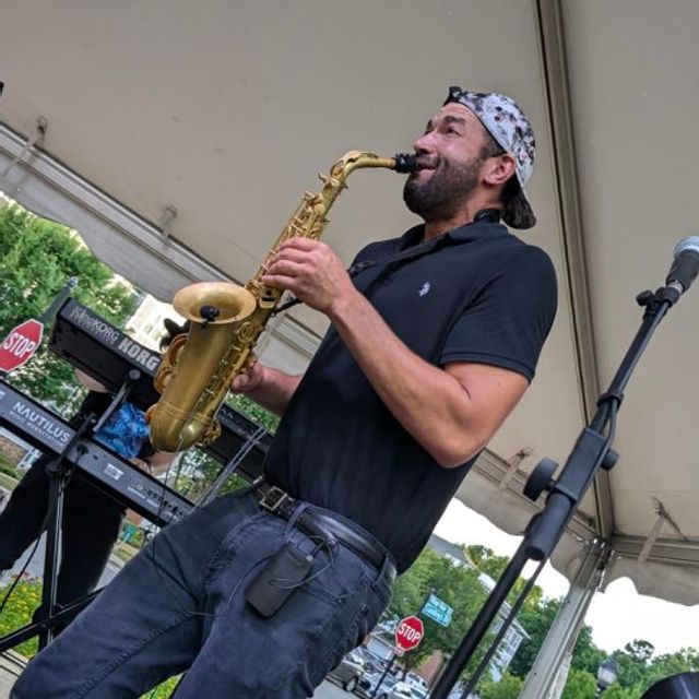 Kory Strother performs onstage while playing a saxophone beneath a white event tent. Dressed in a black polo shirt, jeans, and a patterned backward cap, the performer leans into the music with energy and enthusiasm. Microphones, keyboards, and downtown street signs can be seen in the background, capturing the lively outdoor festival atmosphere.