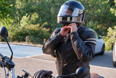 A motorcyclist wearing a black leather jacket fastens the chin strap on a full-face helmet while sitting on a motorcycle in a parking area, with trees and a parked car visible in the background.