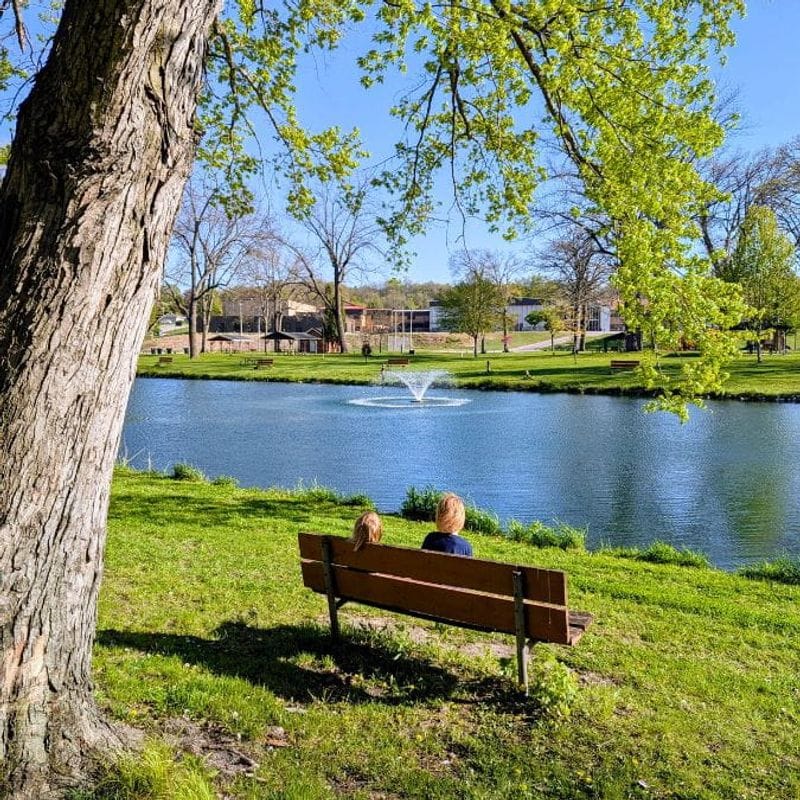 Two young children sit on a bench overlooking a pond and water fountain at Lanesboro's Sylvan Park with a mature tree trunk and leafy branches in the foreground