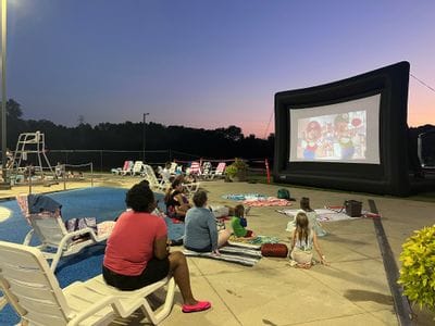 Evening scene at a public pool during a dive-in movie event. Families and children are swimming and playing in the water while a large inflatable movie screen shows a film in the background. A lifeguard stands on the pool deck overseeing the crowd, with bright overhead lights illuminating the area as the sky darkens at dusk.