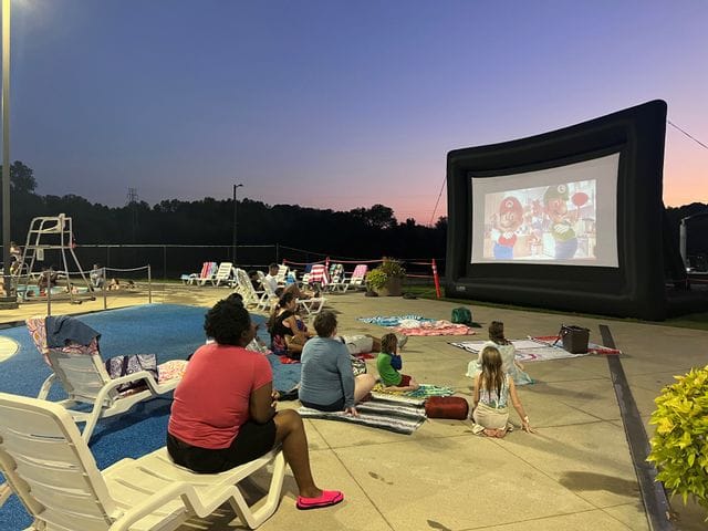 Evening scene at a public pool during a dive-in movie event. Families and children are swimming and playing in the water while a large inflatable movie screen shows a film in the background. A lifeguard stands on the pool deck overseeing the crowd, with bright overhead lights illuminating the area as the sky darkens at dusk.