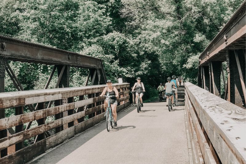 Cyclists ride across a wooden bridge on the Root River State Trail, surrounded by lush green trees on a sunny summer day in Lanesboro, Minnesota.