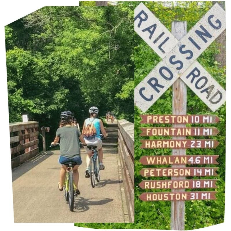 Cyclists ride along the Root River State Trail on a wooded bridge beside a railroad crossing sign listing distances to nearby towns including Whalan, Preston, and Harmony.
