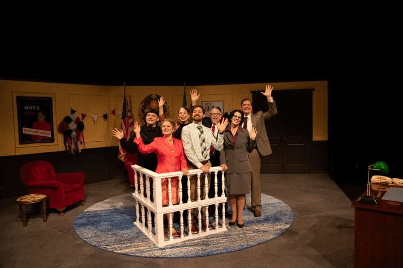 Cast members stand on stage waving to the audience during a curtain call for a Commonweal Theatre Company production, with a detailed set and stage props visible behind them.