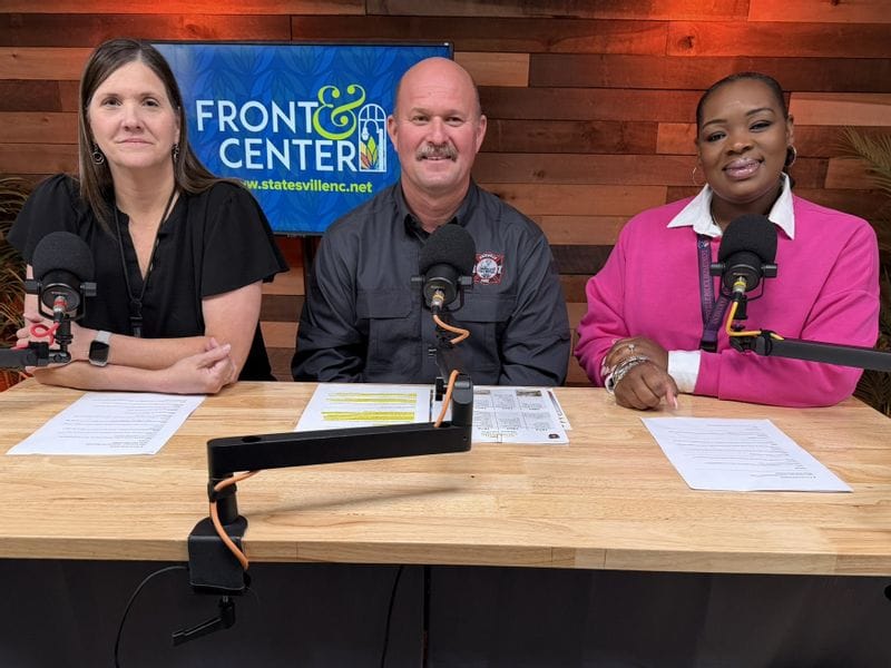 Three podcast hosts sit at a table with microphones in a studio setting. From left to right are April Nesbit, Public Information Officer; Fire Chief Glenn Kurfees of the Statesville Fire Department; and Nakayla Griffin, Community Resource Officer. They are recording an episode of the Front & Center podcast discussing the department’s 150th Anniversary of the Statesville Fire Department.