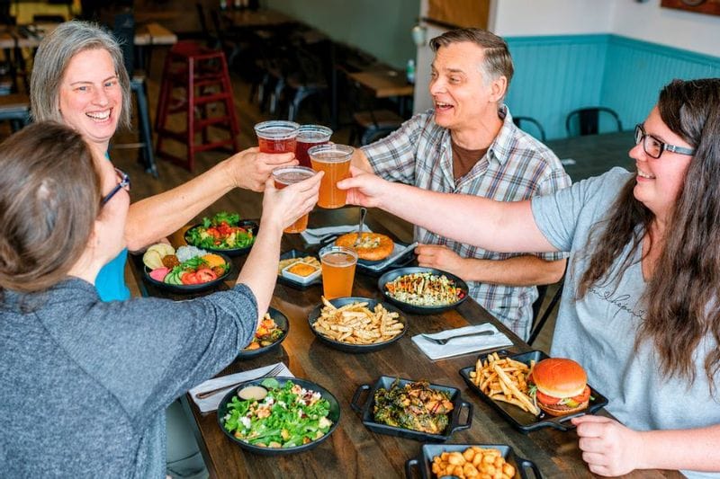 A group of friends raise glasses of beer in a toast around a table at Pedal Pushers Café, surrounded by burgers, fries, salads, and shared plates in a relaxed Lanesboro restaurant setting.