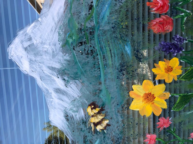 A window painting of a fuzzy bee flying over a bunch of yellow, pink a purple flowers.  In the background is a snow covered mountain.