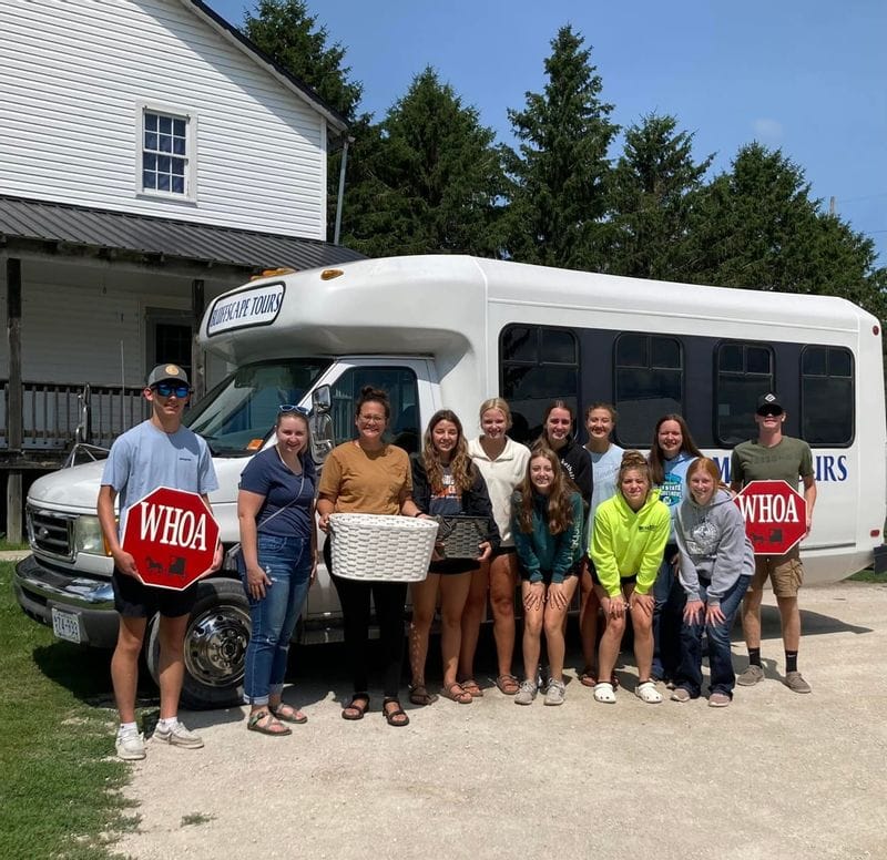Group of visitors posing in front of a Bluffscape Tours shuttle bus outside a rural farm building, with several people holding red “WHOA” buggy stop signs and baskets, standing on a gravel driveway under a clear blue sky with tall evergreen trees behind them.