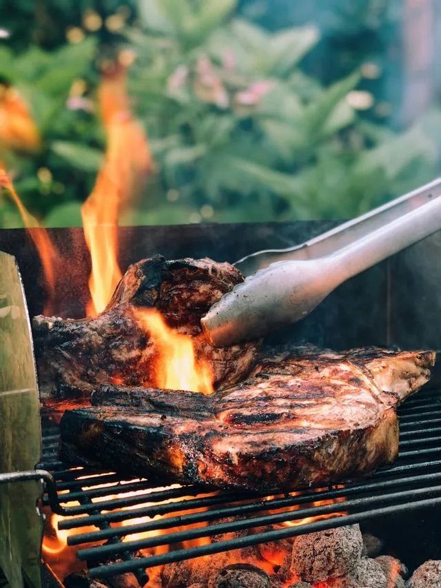 Close-up of steaks cooking on a grill with flames rising as metal tongs flip the meat.