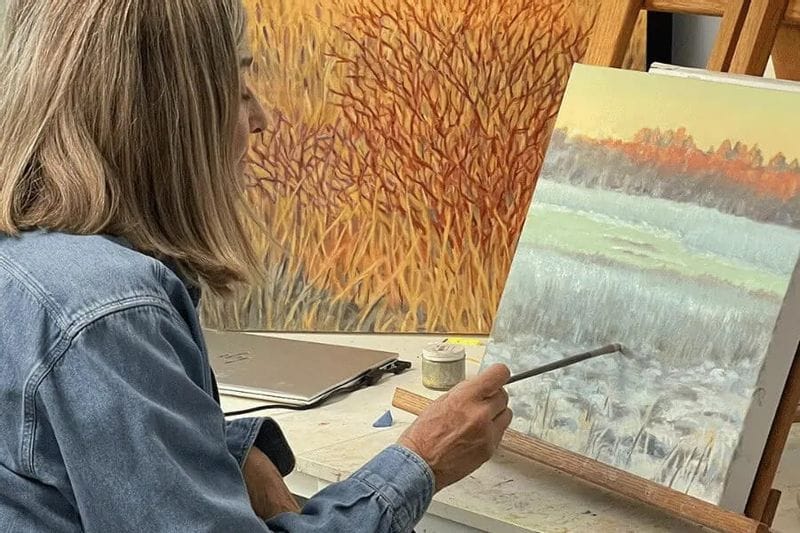 An artist paints a landscape on canvas inside a studio, using a brush to add detail while autumn-colored artwork hangs on the wall behind them.