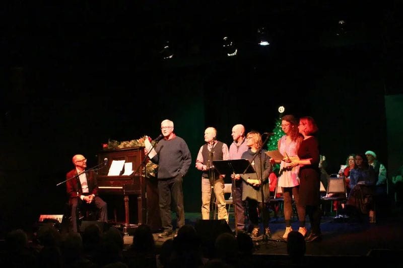 Group of performers and singers on stage during a Lanesboro Arts holiday sing-along, with a pianist at a grand piano and participants holding sheet music under warm stage lighting.