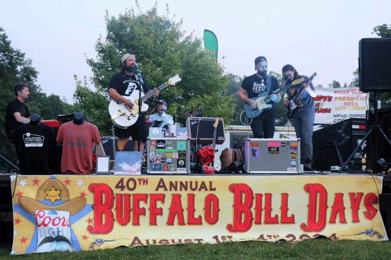 Band performs live on an outdoor stage at Lanesboro’s Buffalo Bill Days, with musicians playing guitars and drums behind a banner reading “40th Annual Buffalo Bill Days,” and festival booths visible in the background.