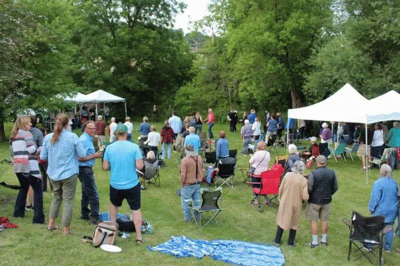 Crowd gathers on a grassy lawn for an outdoor live music event in Lanesboro, Minnesota, with people standing and sitting in lawn chairs near small stages and white canopy tents surrounded by trees.