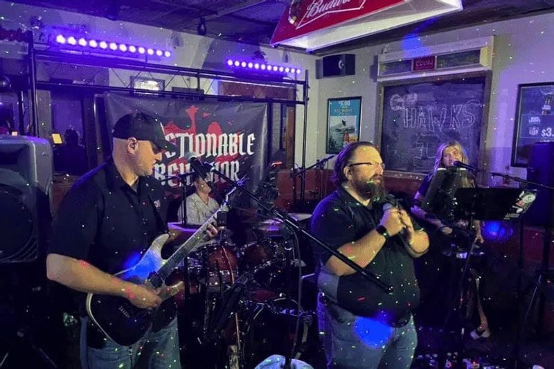 Live band performs on a small stage at the Root River Saloon in Lanesboro, Minnesota, with musicians singing and playing guitar under colorful stage lights in a lively indoor bar setting.