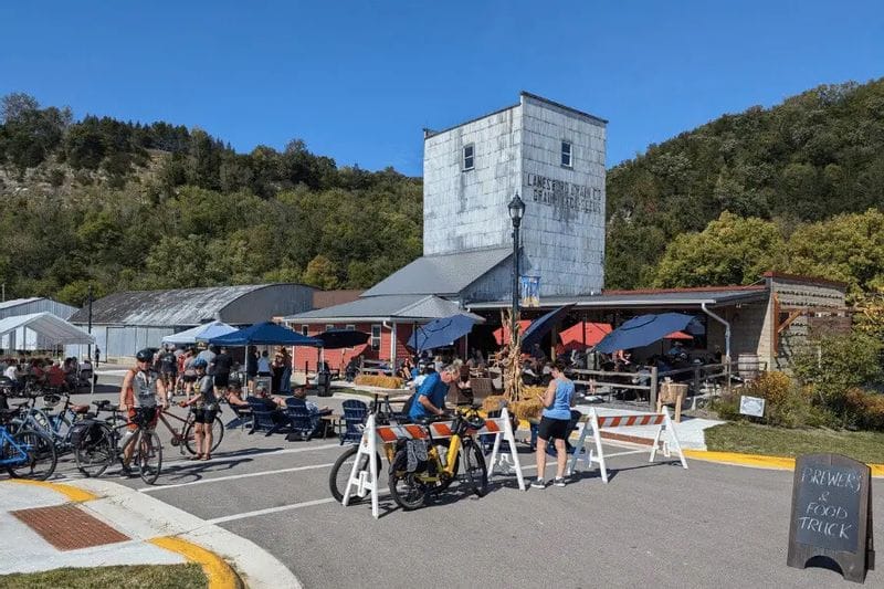 Cyclists and visitors gather outside Sylvan Brewing in Lanesboro, Minnesota, with outdoor seating, live music, and a food truck set up in front of the historic grain elevator building on a sunny day.