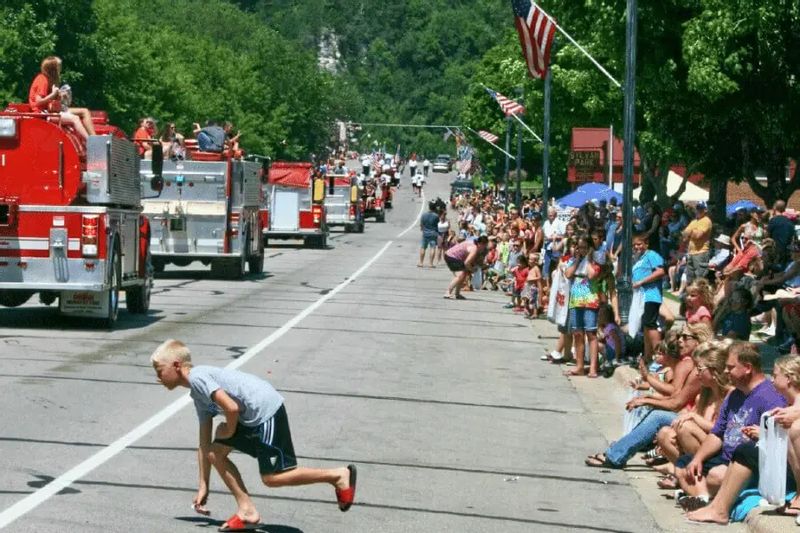 Children and families line the street during a summer parade in Lanesboro as fire trucks drive past, with a young boy running into the road to pick up candy while spectators watch from the curb.